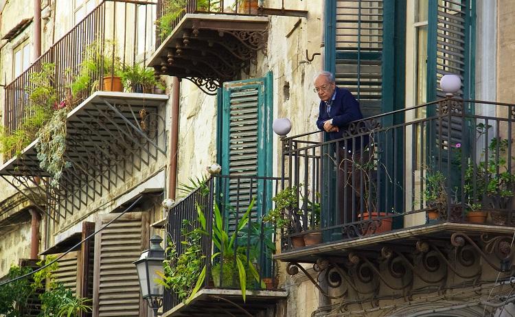 un homme âgé en veste sur son balcon fleuri - mihaly-koles-unsplash