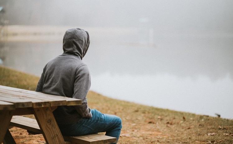 un garçon avec capuche assis sur un banc devant un lac dans le brouillard - amy-tran-unsplash