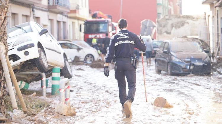 Un policier local de Valence traverse une rue inondée et boueuse suite aux dégâts causés par la DANA. Des voitures endommagées et des bâtiments touchés sont visibles en arrière-plan.