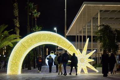 Un décor féerique à Barcelone pour Noël 2024 : une arche lumineuse en forme d'étoile dorée éclaire une promenade bordée de palmiers, invitant les visiteurs à profiter de la magie des fêtes