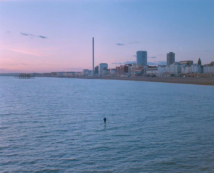 Un homme sur son paddle dans la mer, à l'arrière-plan une vue de la ville en début de soirée 